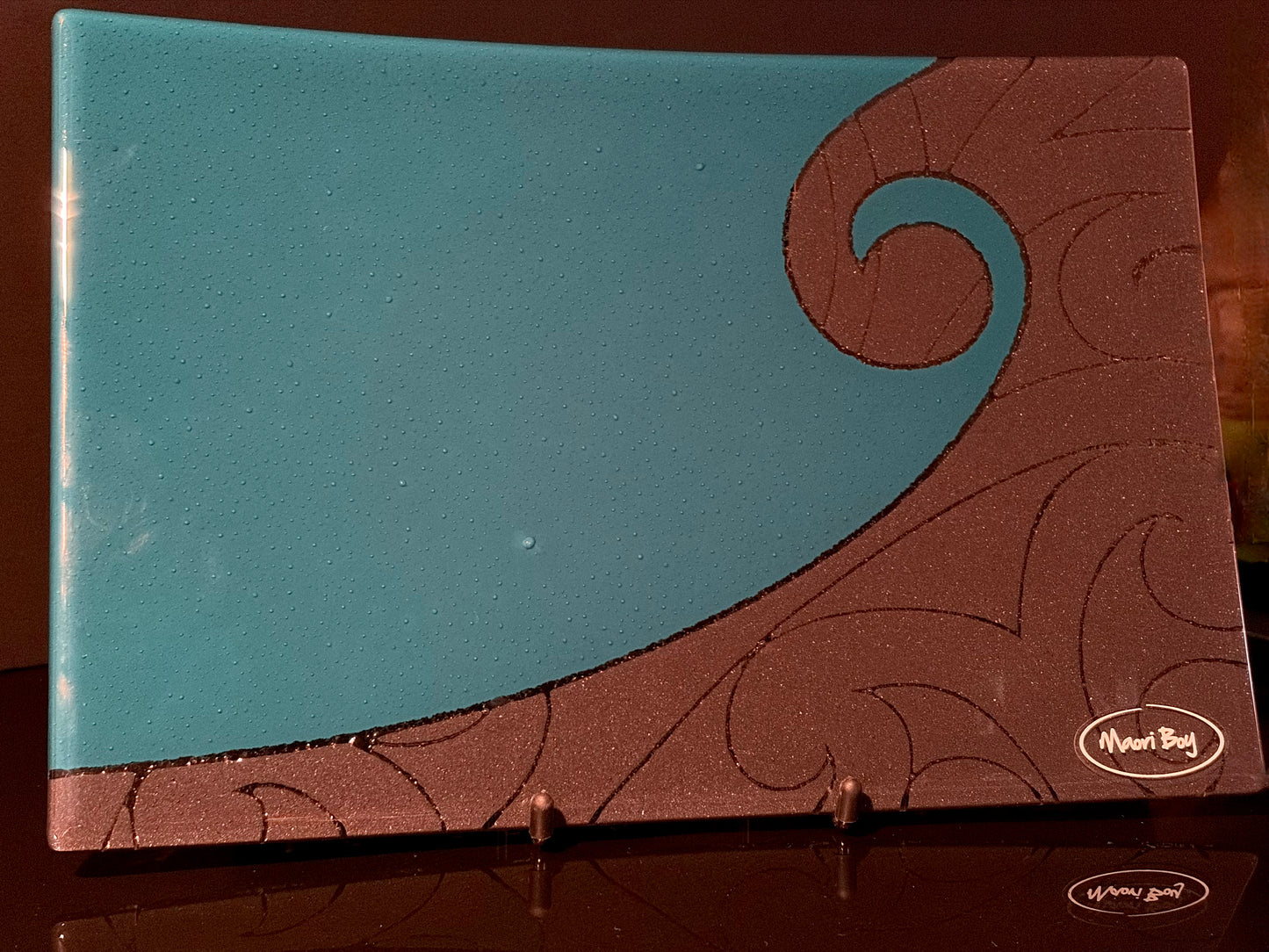 Decorative plate with teal and black Maori koru design on a reflective surface.