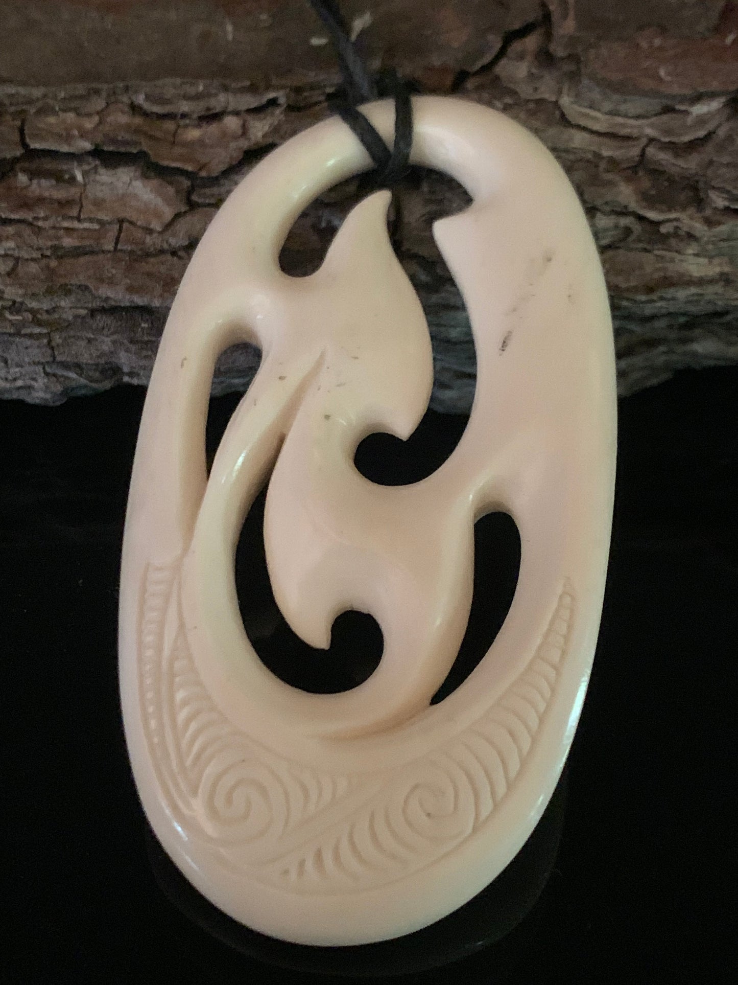 Reverse side of a bone freeform pendant with traditional Maori carvings, suspended on a waxed nylon cord against a wooden background.