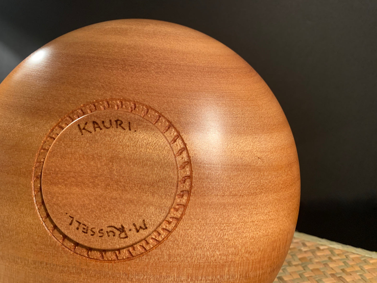 Wooden bowl base with 'Kauri' and artists signature engraving on a dark background.
