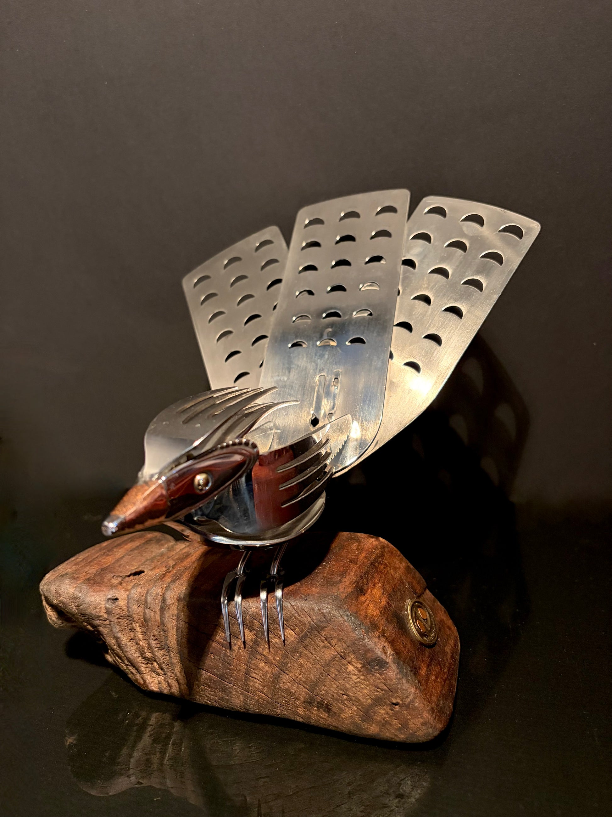Metallic sculpture of a New Zealand fantail bird on a wooden base.  Made from kitchen utensils.