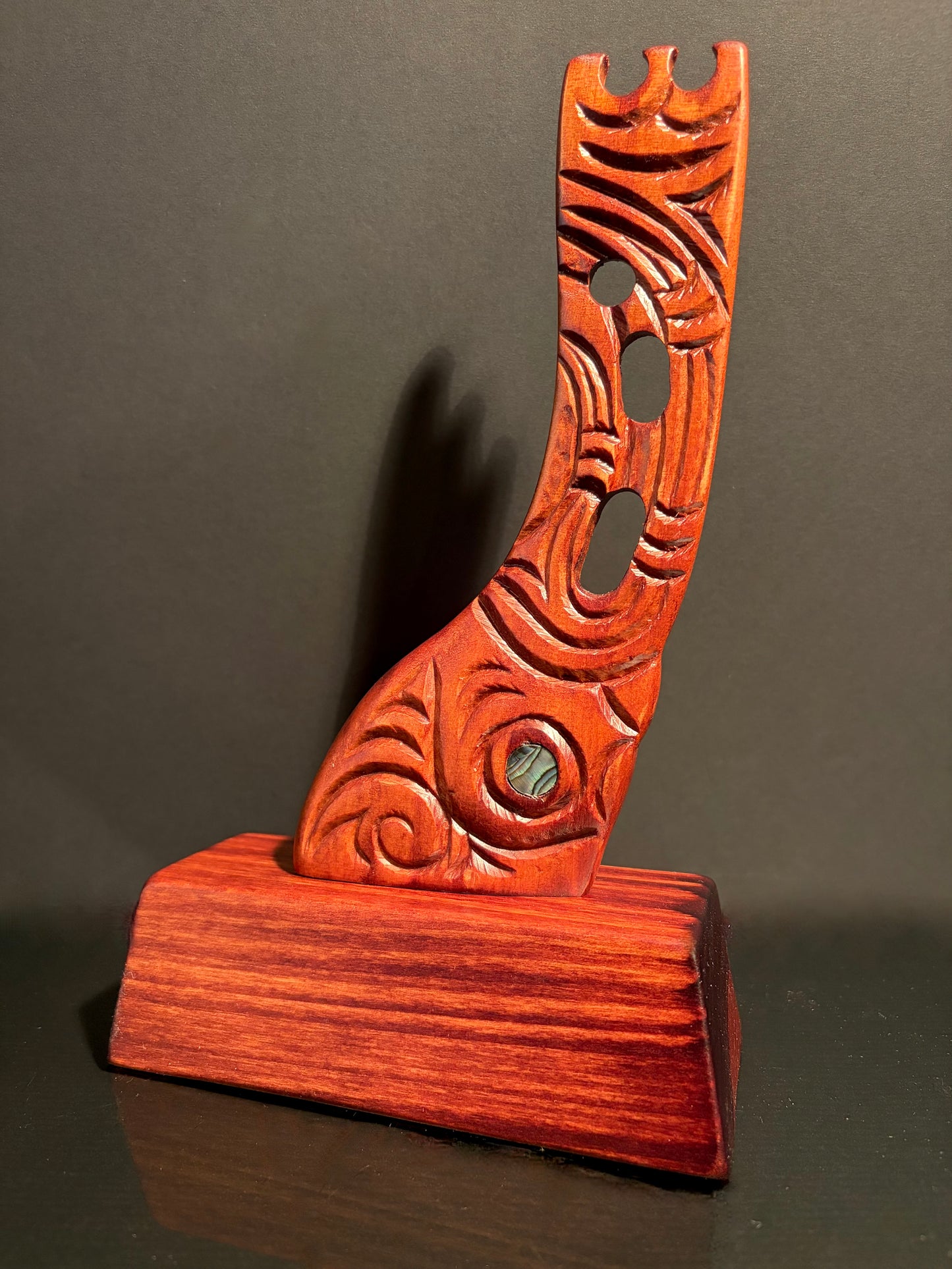 Wooden carved Maori waka (canoe) stern carving with intricate designs and paua detail on a wood stand.