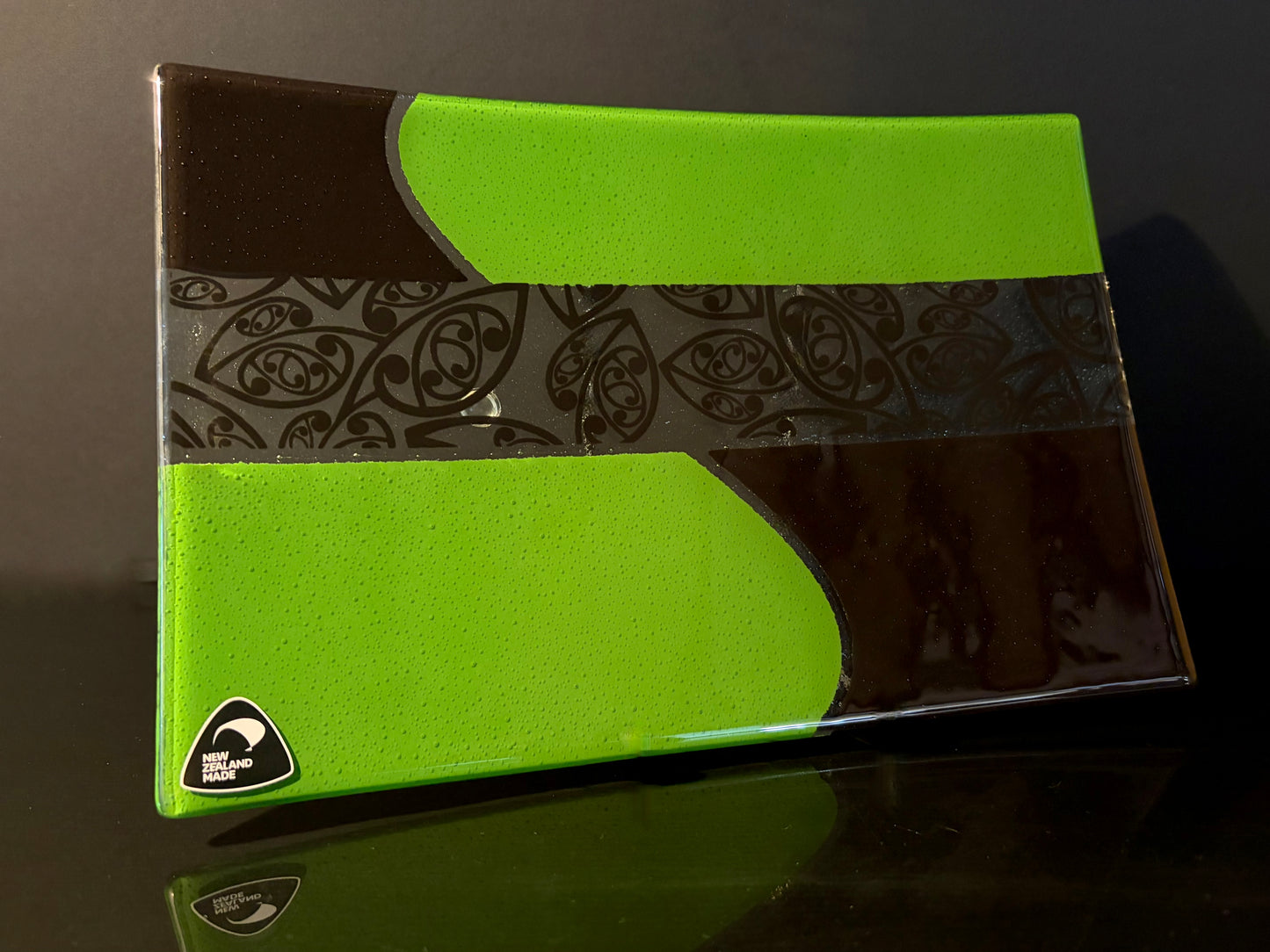 angled view of green and black rectangular fused glass plate with maori kowhaiwhai pattern on a dark background