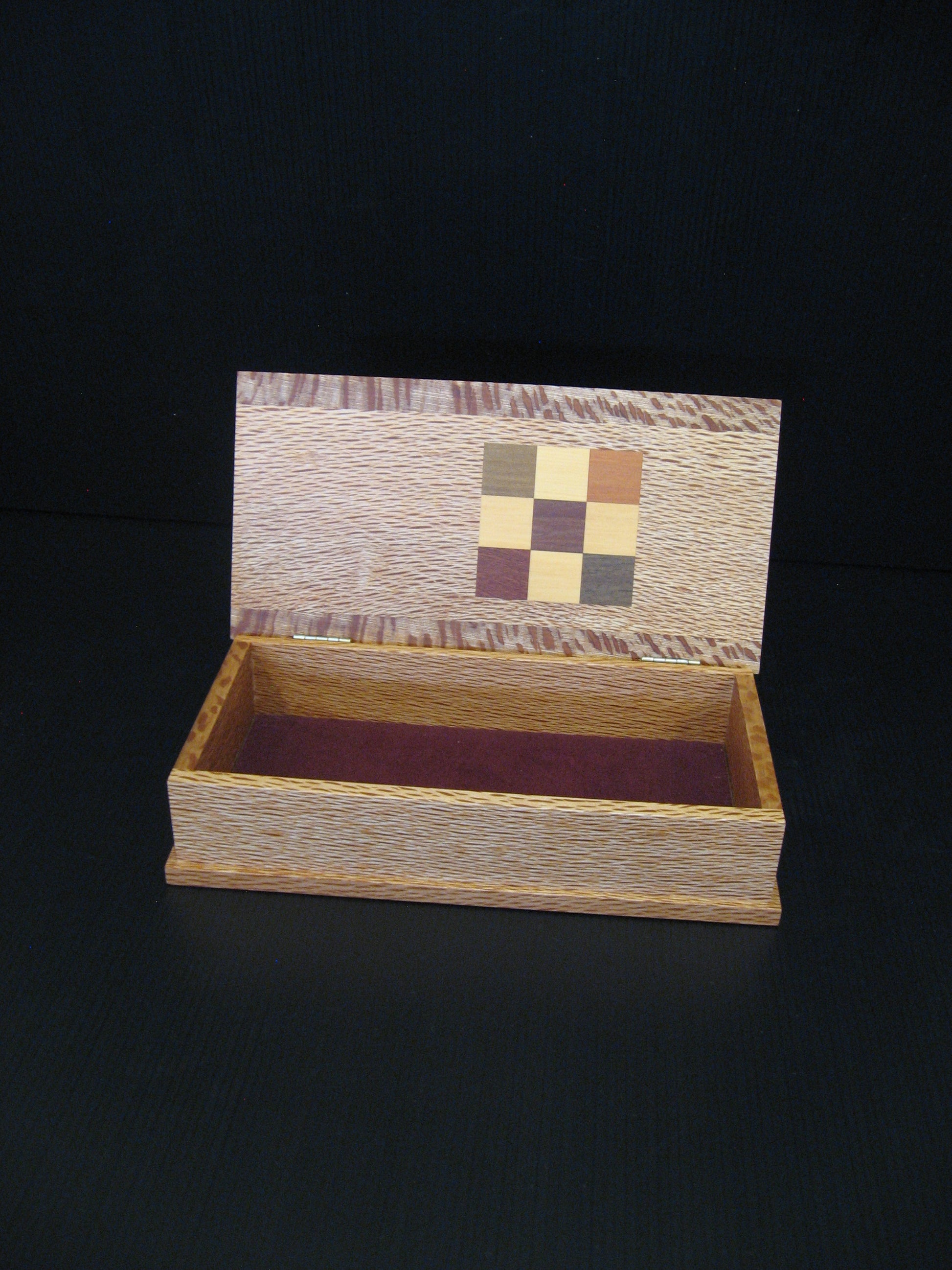 A small open wooden keepsake box with a hinged lid, featuring a decorative multicolor checkerboard inlay on the inside of the lid and a dark burgundy velvet lining.