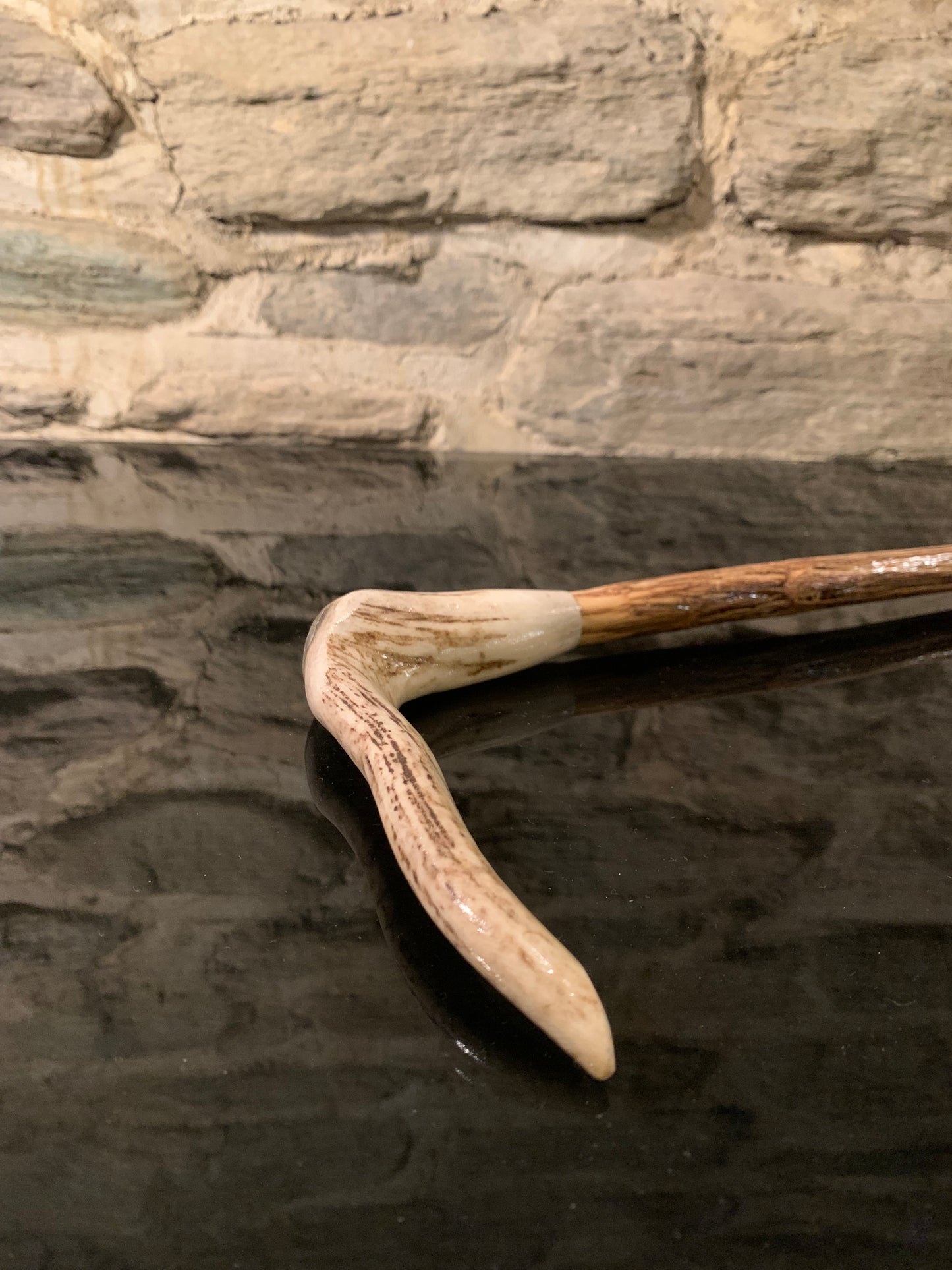 A curved deer antler walking stick with a brown wooden stick displayed against a textured stone wall
