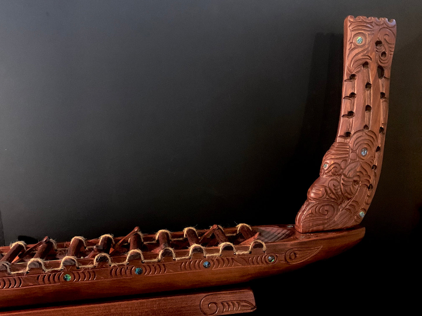 An intricately carved wooden Maori waka (canoe) with detailed patterns and small blue-green NZ paua shell accents