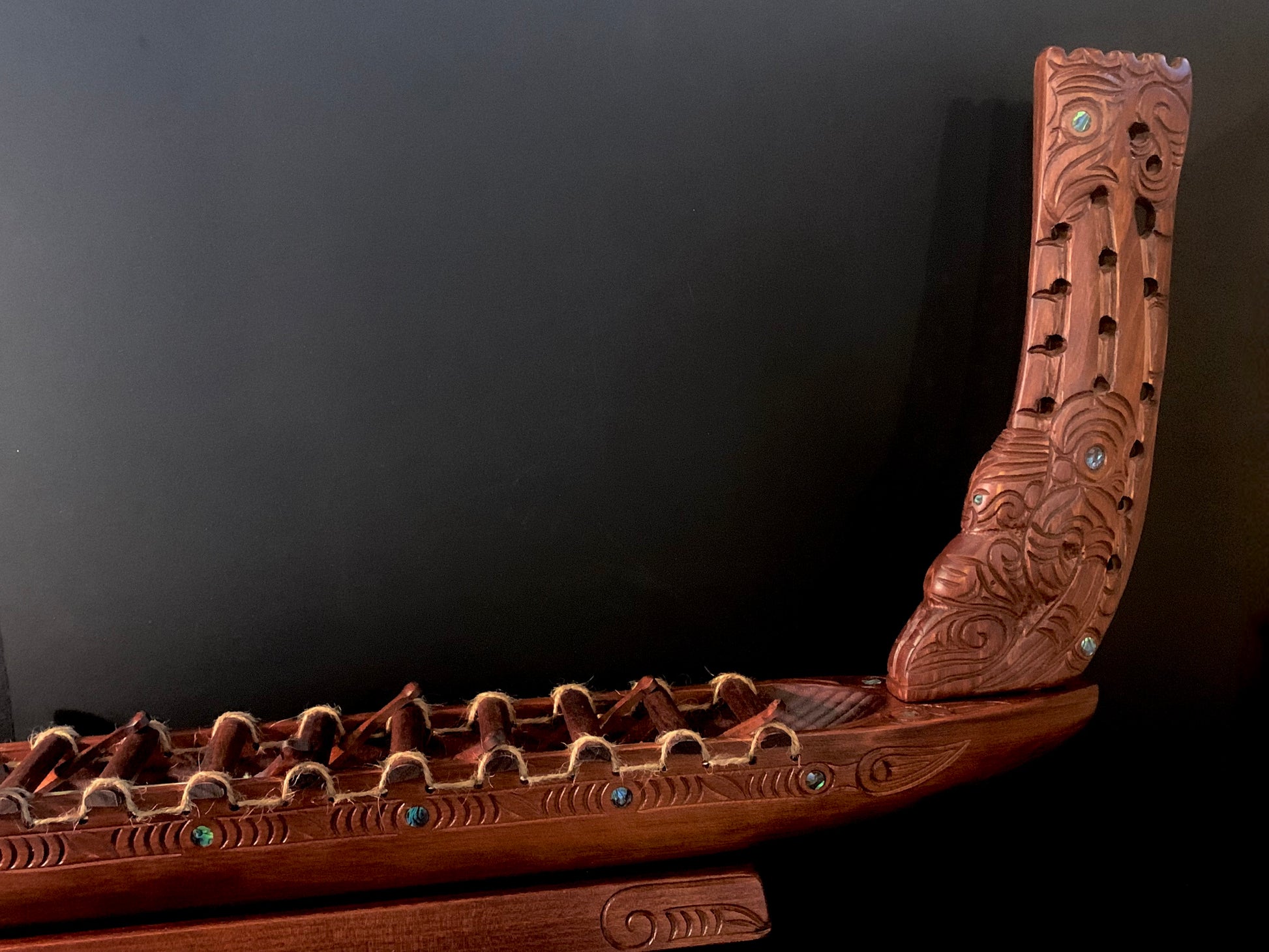 An intricately carved wooden Maori waka (canoe) with detailed patterns and small blue-green NZ paua shell accents
