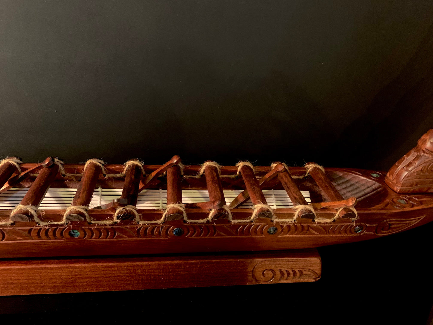 A close-up of a carved wooden Maori waka (canoe) with brown wood, tan rope, and small blue paua shell accents